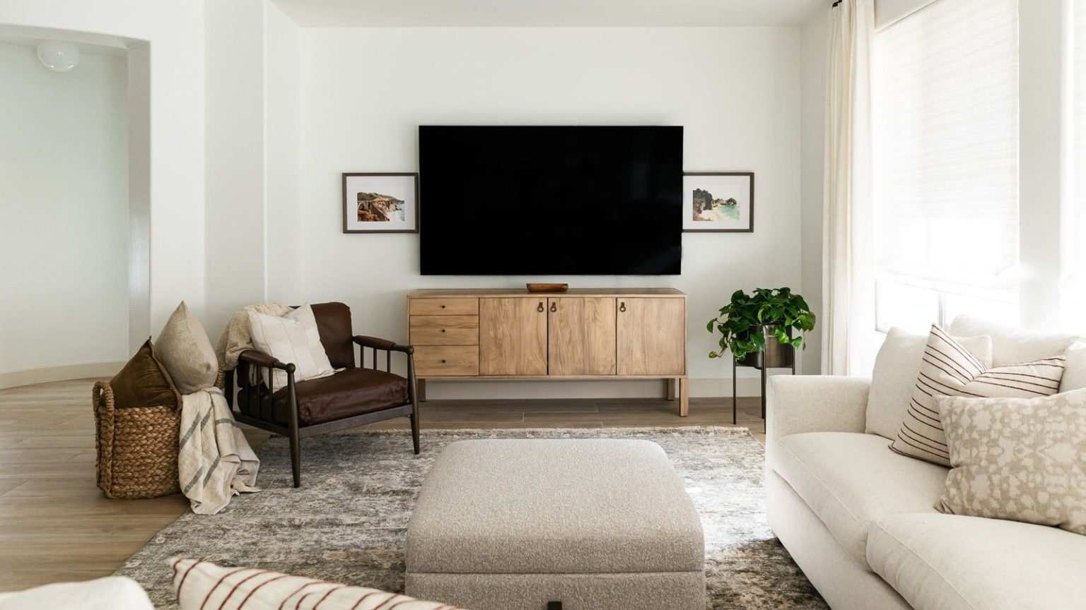 La barre de son Sonos Arc Ultra offre des détails magistraux et un son immersif – maintenant avec une rare réduction de 200 $ Living room with TV mounted on wall above retro wooden media unit, with cream sofa and footstool pictured in foreground. Also pictured are a brown armchair, miscellaneous cushions, a potted plant, and two framed pictures, in a room with white walls and curtains.