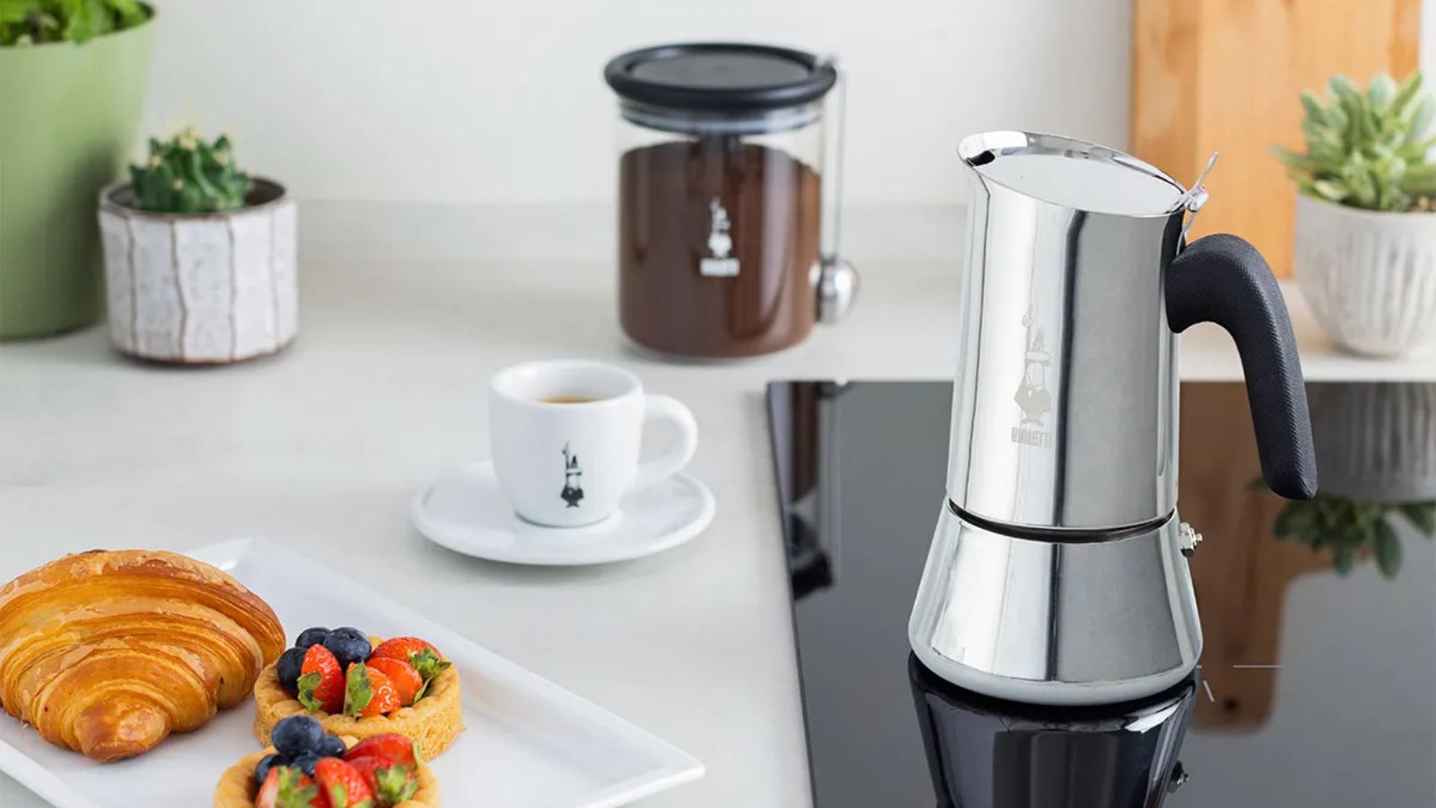 Une cafetière Bialetti argentée sur une cuisinière électrique noire, installée dans un comptoir blanc. À côté, une petite tasse à expresso, avec une assiette carrée composée d'un croissant et de quelques flans aux fruits frais. 