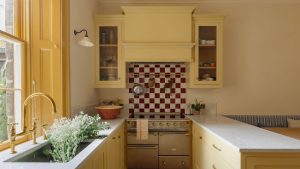 Vous ne trouverez même pas une touche de bleu dans cette petite cuisine côtière. Voici pourquoi les concepteurs ont utilisé une teinte beurrée inattendue pour aller à l’encontre des clichés de couleurs dépassés. Horseshoe layout kitchen with yellow Shaker cabinets, brass hardware, red and white checkerboard backsplash, farmhouse sink, and built-in banquette seating.