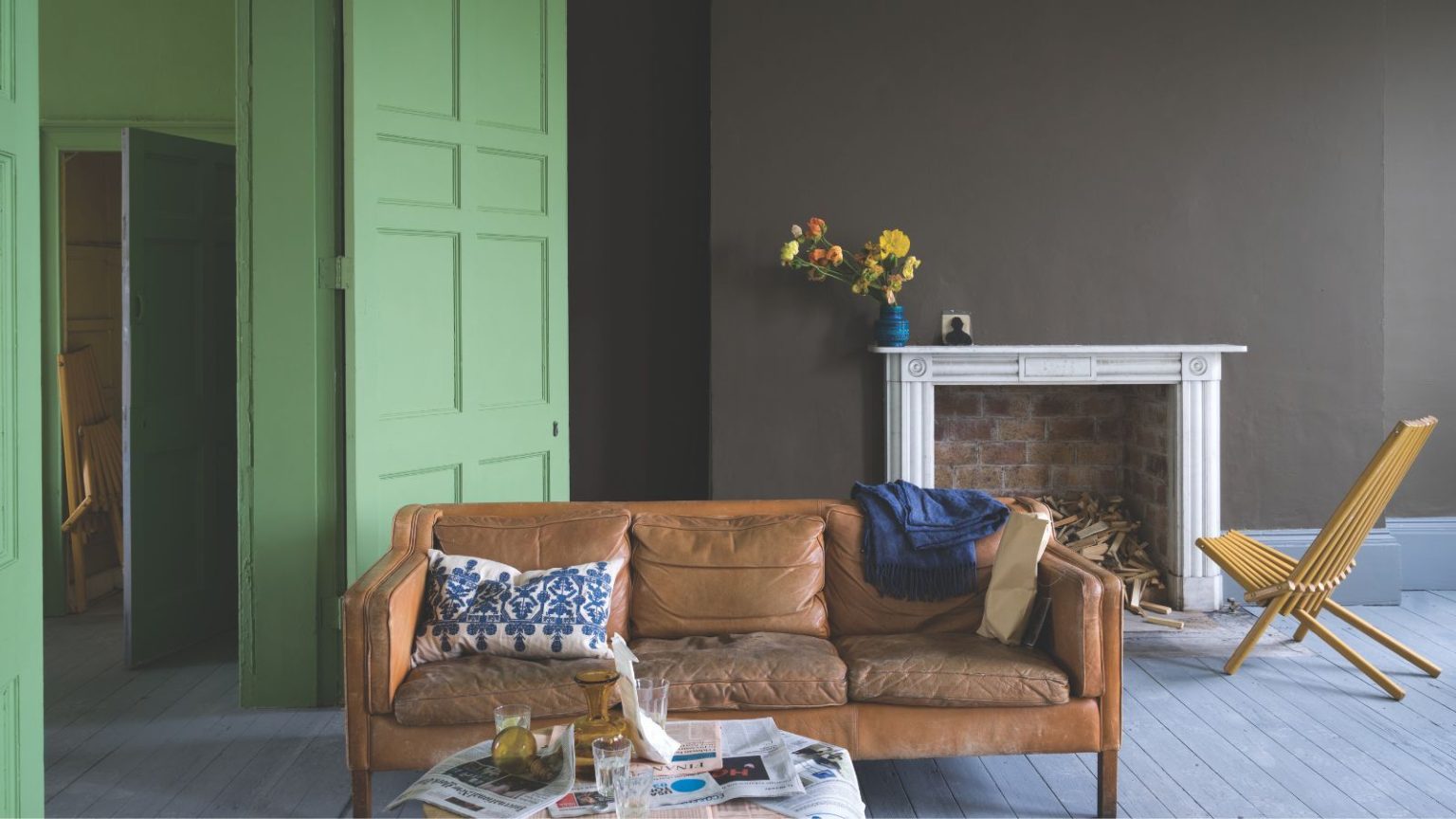 « Un brun riche et terreux qui change doucement avec la lumière » – Pourquoi les designers se tournent toujours vers cette couleur Farrow & Ball « Caméléon » pour un confort instantané Living room with wooden floorboards, central wall painted dark brown around the large fireplace, a leather couch at the centre of the room, with a lime green painted door and hallway