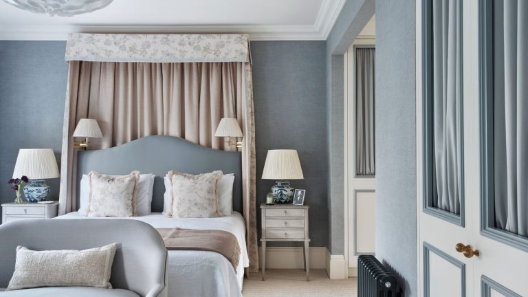 Bedroom with pale blue walls, a floral canopy and pale blue headboard, neutral bedding, white vintage nightstands, and a small blue loveseat