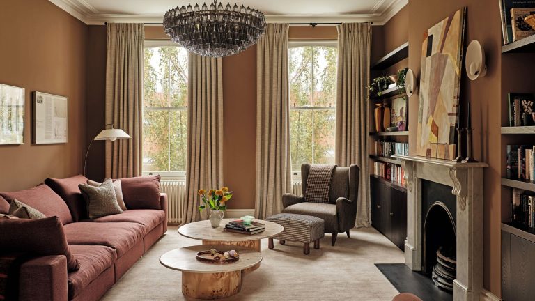À l’intérieur d’une maison de ville londonienne tranquille et élégante, transformée avec des couleurs chaudes et des courbes apaisantes A warm, earth-toned living room featuring terracotta walls, a dusty rose sectional sofa, and a large tiered glass chandelier. Two organic-shaped wooden coffee tables sit on a plush beige rug before a marble fireplace.