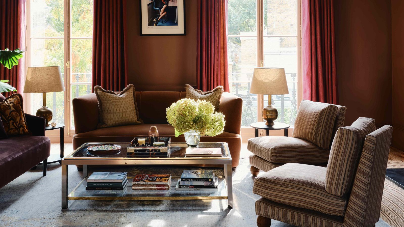 Salon marron orange foncé avec des rideaux orange, de grandes fenêtres, un canapé beige, des fauteuils à rayures avec des pieds en chignon et une table basse en verre