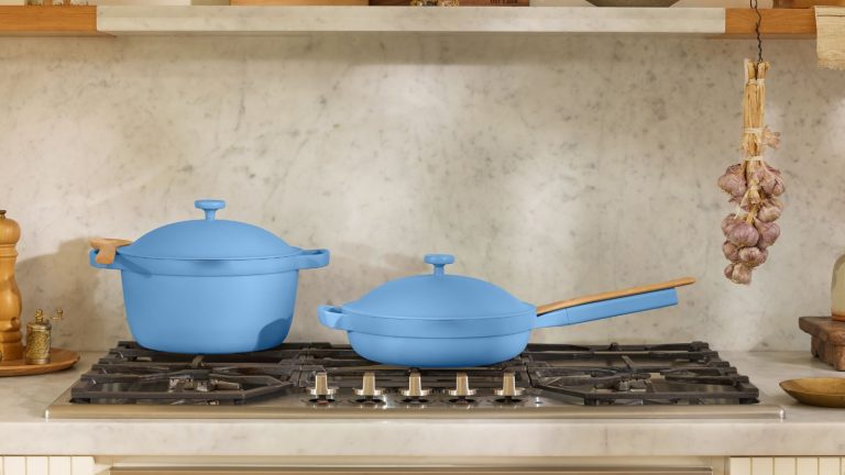 Le Do-Everything Our Place Always Pan est désormais disponible en bleu poudre – et nous prévoyons que ce best-seller non toxique sera rapidement en rupture de stock A blue Dutch oven pan, and a matching blue frying pan with lids on a black gas burning stove top. There is a matching greige stone backsplash and countertop, and garlic bulbs hand from a shelve above.