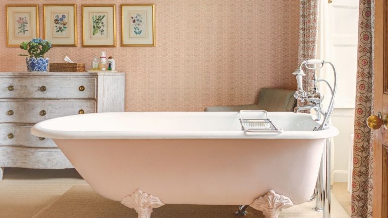 Adieu les tapis de bain humides – Cette mise à niveau en pierre à séchage rapide est la solution de luxe pour salle de bain dont je ne savais pas avoir besoin A pink wallpapered bathroom with a rolltop free standing tub. A distressed dresser in the background with eight gold framed pictures on the wall above it.