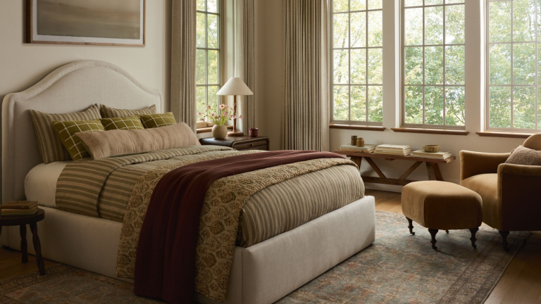 Joon Loloi vient de lancer sa première collection de chambres à coucher – et elle est aussi raffinée, superposée et intemporelle que vous l’imaginez a bedroom with neutral colors, an upholstered bedframe with green beddings, an accent chair with a small ottoman, and a wall of windows with curtains hanging in front