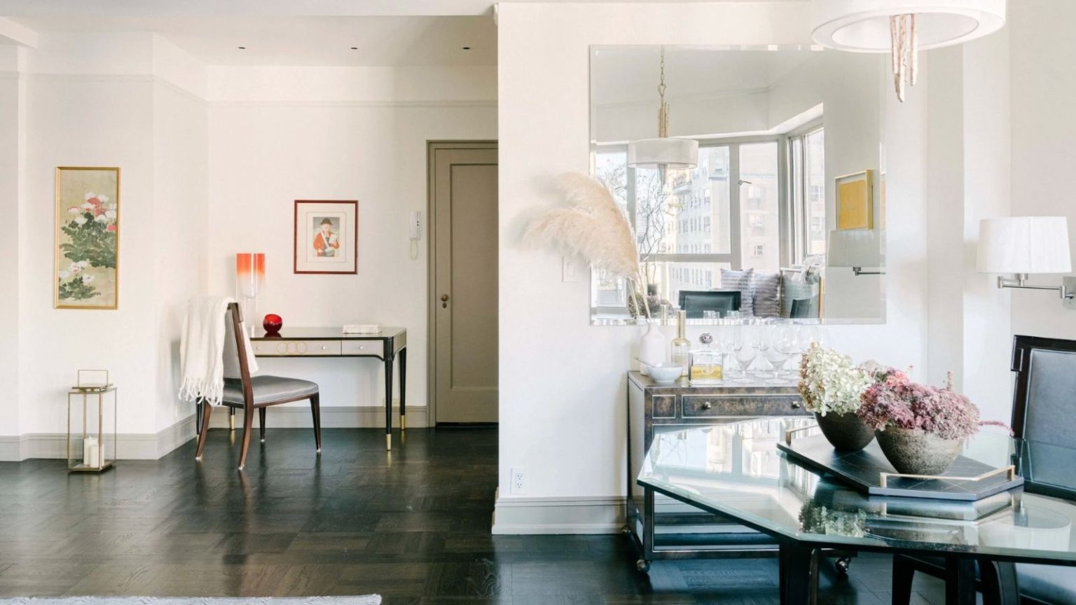 Bright living space with glass tables, chairs, white walls, framed art, a large mirror, and a wooden floor.