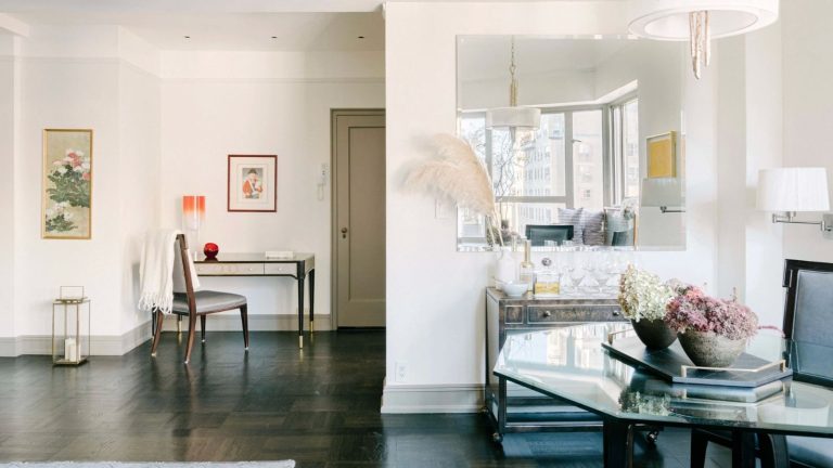 Bright living space with glass tables, chairs, white walls, framed art, a large mirror, and a wooden floor.
