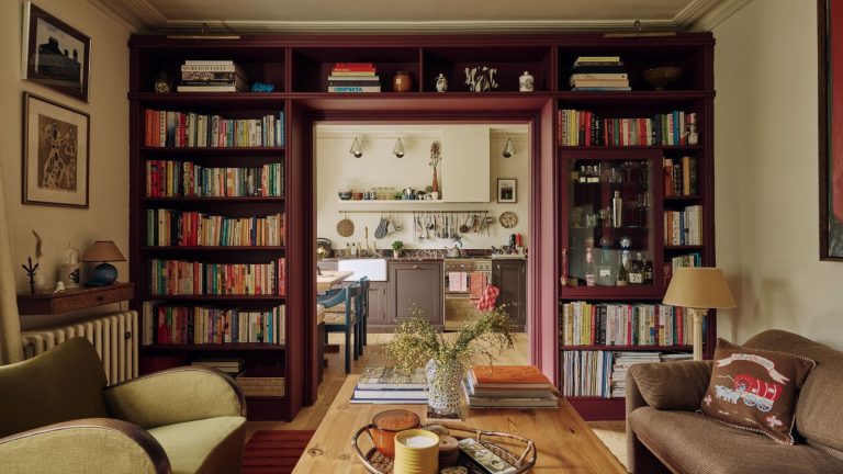 Les designers ajoutent des « coins intimes » aux salons – voici pourquoi ils se sentent si luxueux Living room with a built-in bookcase surrounding the doorway, painted burgundy, filled with books and a built-in bar. The living room features a wooden coffee table with candles, a vase, and books, a velvet green armchair, a brown sofa and looks through onto the kitchen