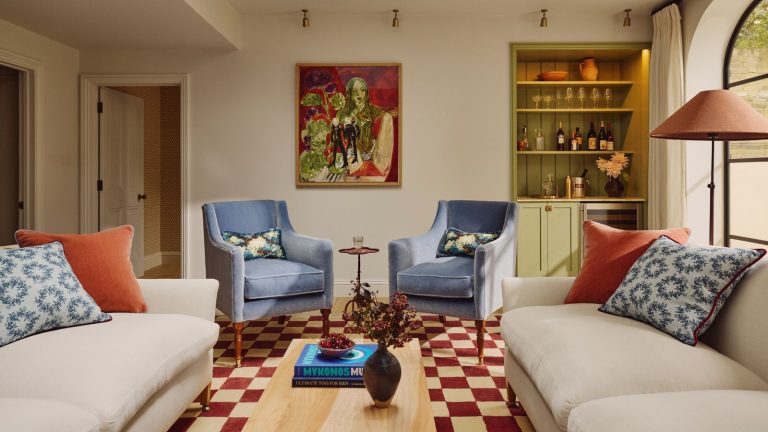 Je vous le promets, cette couleur de canapé peut donner l’impression que n’importe quel petit salon est deux fois plus grand – et les designers sont d’accord neutral living room with bold art, two white sofas, blue armchairs, red checkerboard rug and a green home bar in the corner