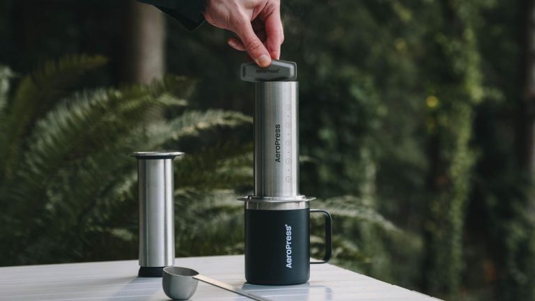 Le nouvel AeroPress en acier inoxydable est la mise à niveau définitive pour l’amateur de café soucieux du design A stainless steel AeroPress tool on top of a black travel mug being held in place by someone's fingers on top, on a white table. Green leafy plants out of focus in the background.