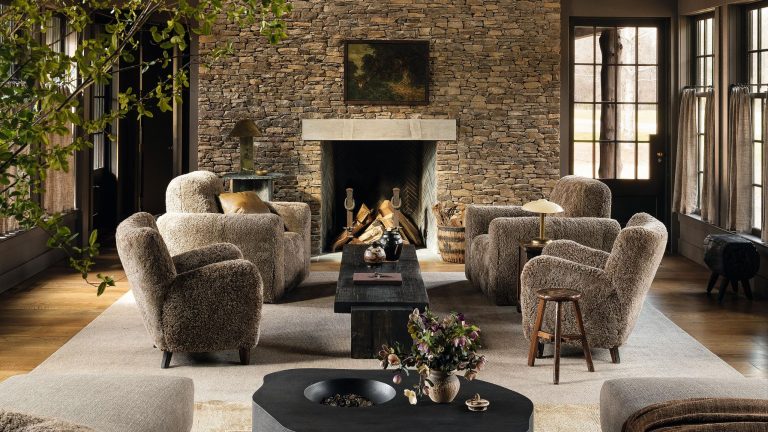À l’intérieur d’une ferme maussade de l’Alabama cachée parmi les arbres, où la cape du cocher de Benjamin Moore occupe le devant de la scène – « Nous avons choisi les couleurs que nous voyions dehors » a moody modern farmhouse living room with a large stone fireplace and four sheepskin armchairs styled by the fire on a large rug with dark black wooden rustic tables