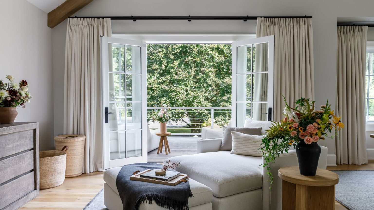 L’édition très britannique : Comment amener l’extérieur à l’intérieur peut transformer votre maison – Créez des pièces plus lumineuses, plus légères et pleines de vie Large living area with open balcony window finished with cream curtains, view outdoors to blurred greenery. There is a cream daybed in the center of the room with a wooden side table decorated with a vase of flowers placed to the right, a wooden cabinet, and neutral baskets to the left-hand side, a gray rug to the right.