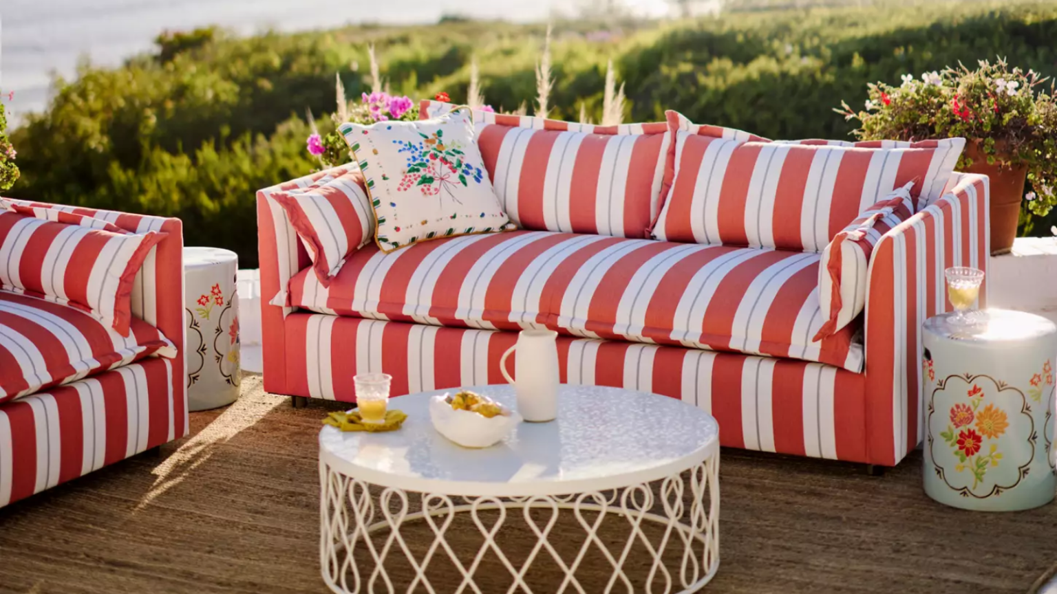 Bye Bye les meubles d’extérieur ennuyeux, le patio retrouve sa personnalité avec la nouvelle version d’Anthropologie a red and white striped outdoor couch with a pillow on it, a white outdoor coffee table with decorative detailing around the sides, a jute rug, and a small accent table to the right of the couch