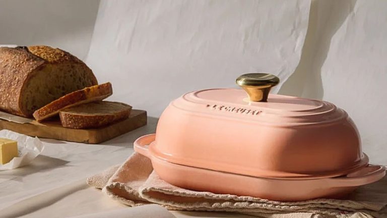 Arrêtez de vous contenter du pain moyen : le nouveau four à pain ovale Signature de Le Creuset apporte des pâtisseries artisanales directement dans votre cuisine A pink cast iron oval bread oven on a folded dish towel, with a loaf of fresh sliced bread in the background.