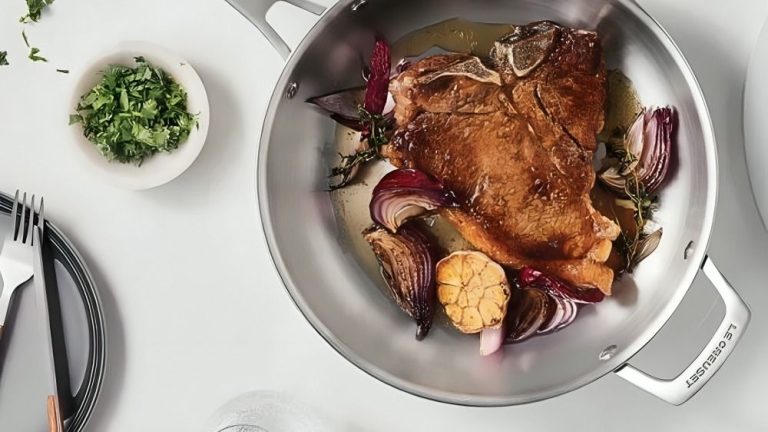 J’avais trop peur pour essayer une poêle en acier inoxydable, mais j’admets que je me trompais : c’est le meilleur interrupteur de cuisine non toxique que j’ai fabriqué en 7 ans. A large stainless steel frying pan with a carry handle on a white counter. A fried steak inside with red onion and a garlic bulb. Beside it is a small dish of chopped parsley, and a plate with a knife and fork.