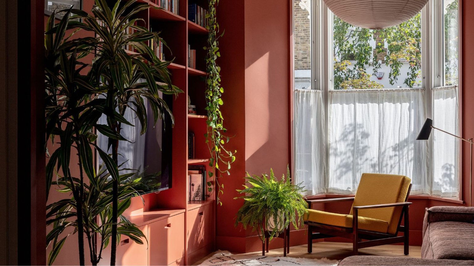 Adieu le rouge cerise, le rouge terreux donne à votre maison une sensation beaucoup plus ancrée – et chaque pièce donne l’impression d’être dans une heure d’or permanente. Minimal living room with earthy red painted walls and a built-in bookcase with TV, a lantern pendant light, a yellow midcentury chair, a brown boxy sofa, a Berber rug and a gold floor lamp in the corner beside a large bay window with cafe curtains and lots of large potted plants