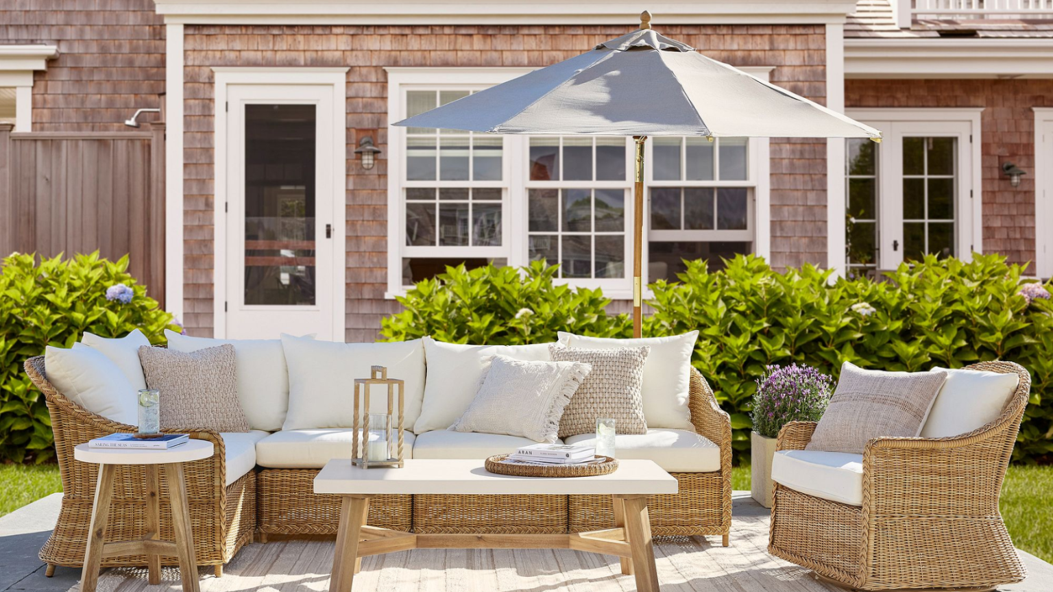 Aucun patio chic n’est complet sans un tapis d’extérieur – Ces 24 choix donneront enfin à votre jardin l’impression d’être un salon a Cape Cod style outdoor patio with wicker furniture and white cushions, a grey outdoor umbrella, and a cedar shingled house