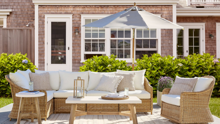 a Cape Cod style outdoor patio with wicker furniture and white cushions, a grey outdoor umbrella, and a cedar shingled house