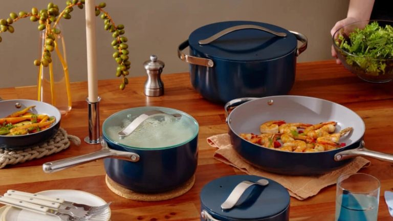 A set of dark blue ceramic non-stick pans on a wooden dining table. A sauce pan with a glass lid, a frying pan filled with fried shrimp, and a larger stock pot with a matching blue lid.