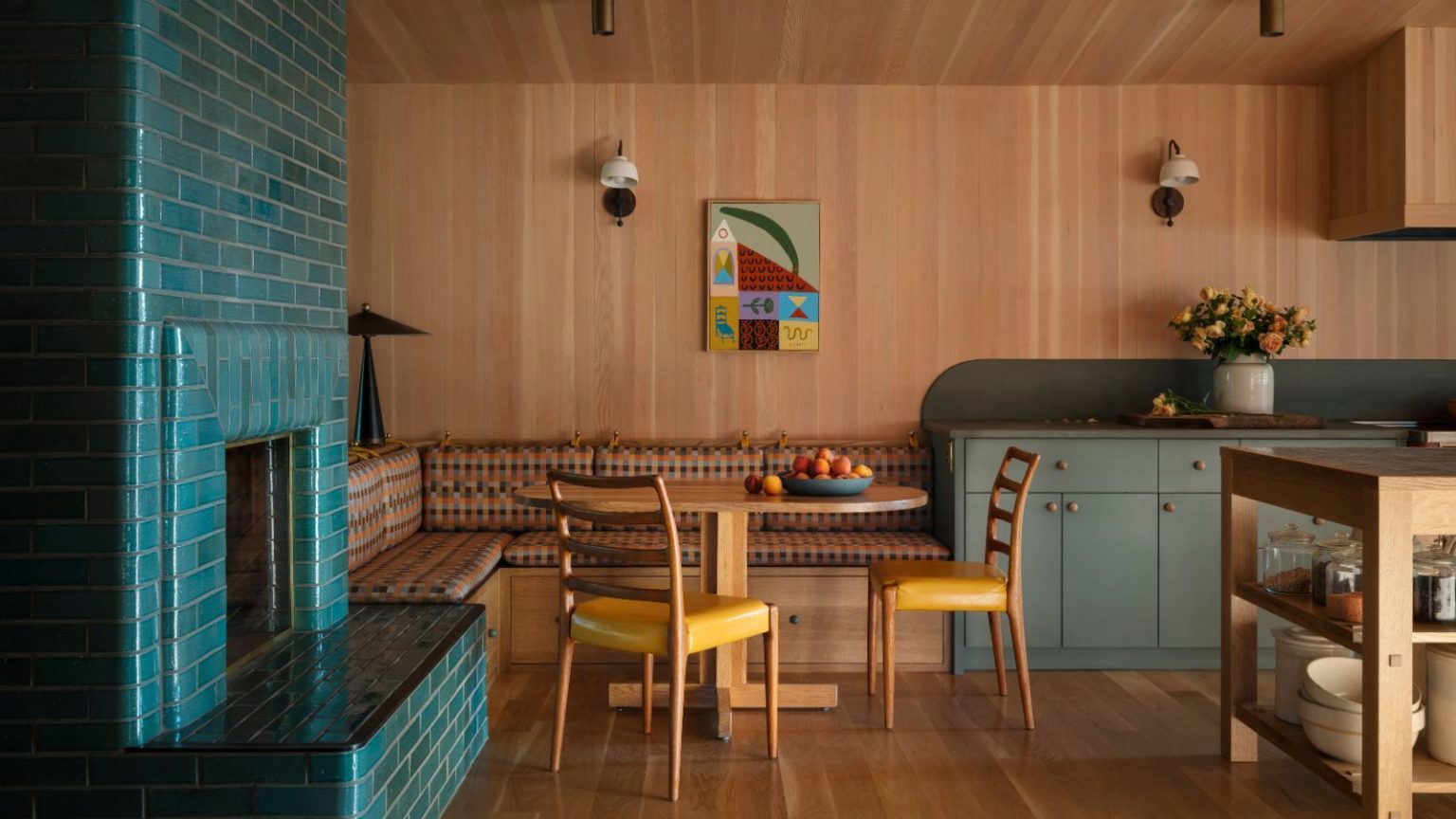 Comment les designers décorent avec des couleurs nostalgiques inspirées des années 70 d’une manière qui semble moderne et fraîche, pas démodée A wood-clad open-plan kitchen with wood floors, gray-green cabinets, banquette seating with a wood dining table and two mustard chairs. A teal brick fireplace.