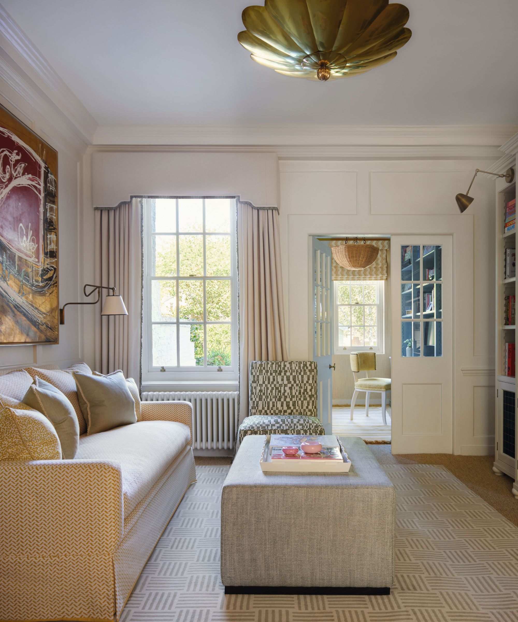 Un salon confortable avec un canapé à motifs crème, un grand pouf gris et un plafonnier festonné doré. Une porte mène à un bureau à domicile ensoleillé avec une chaise jaune.