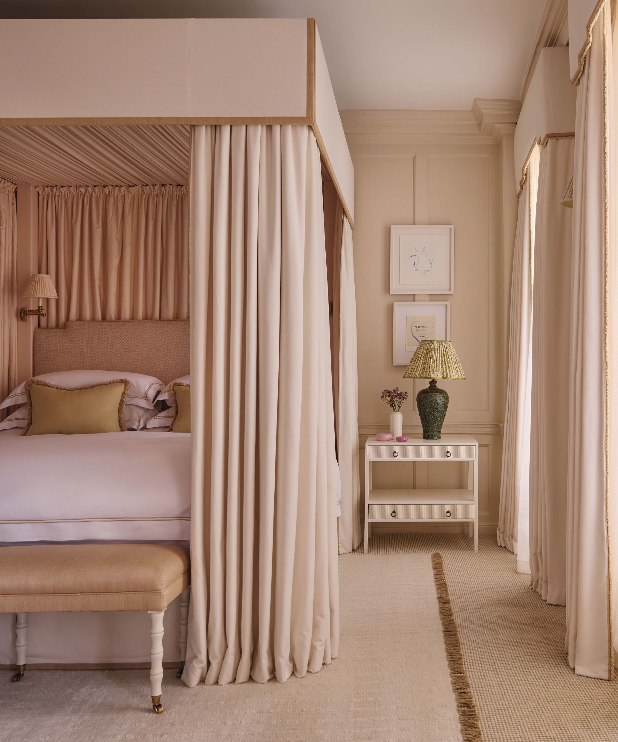 Une chambre sereine et monochrome dotée d'un grand lit à baldaquin drapé d'un épais tissu crème. La pièce est décorée avec une table de nuit crème assortie, une lampe en céramique verte avec un abat-jour plissé et des lambris muraux neutres et doux.