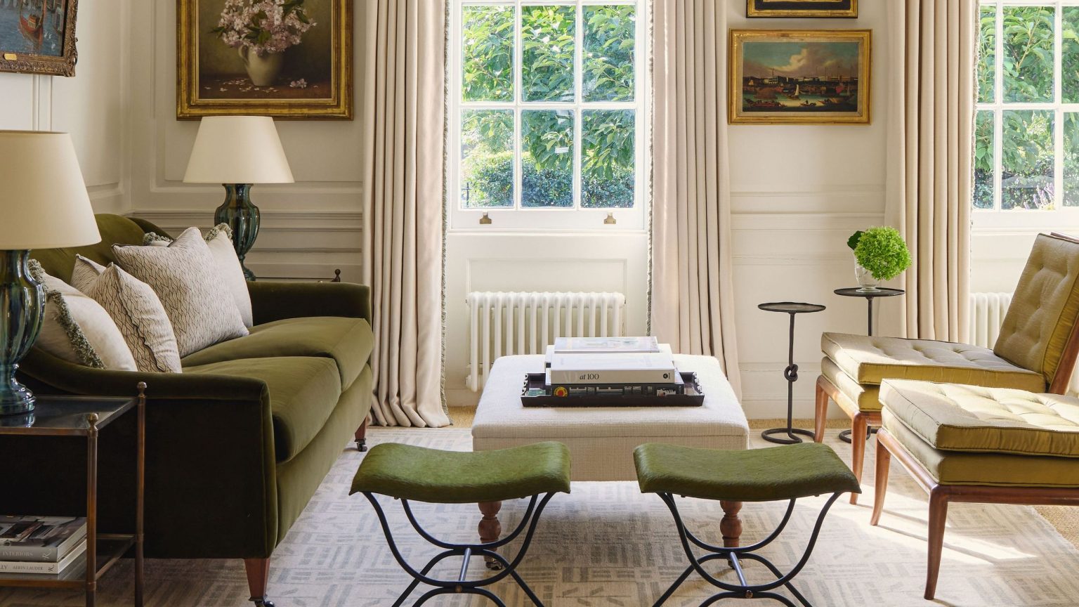 Cette maison londonienne tranquillement confiante cache un intérieur étonnamment ludique – « Tout était si simple qu’il fallait quelque chose d’inattendu » A bright, traditional living room featuring a forest green velvet sofa, olive green mid-century chairs, and twin green stools. Large sash windows are framed by cream drapes and a formal pelmet, with classical oil paintings on the walls.
