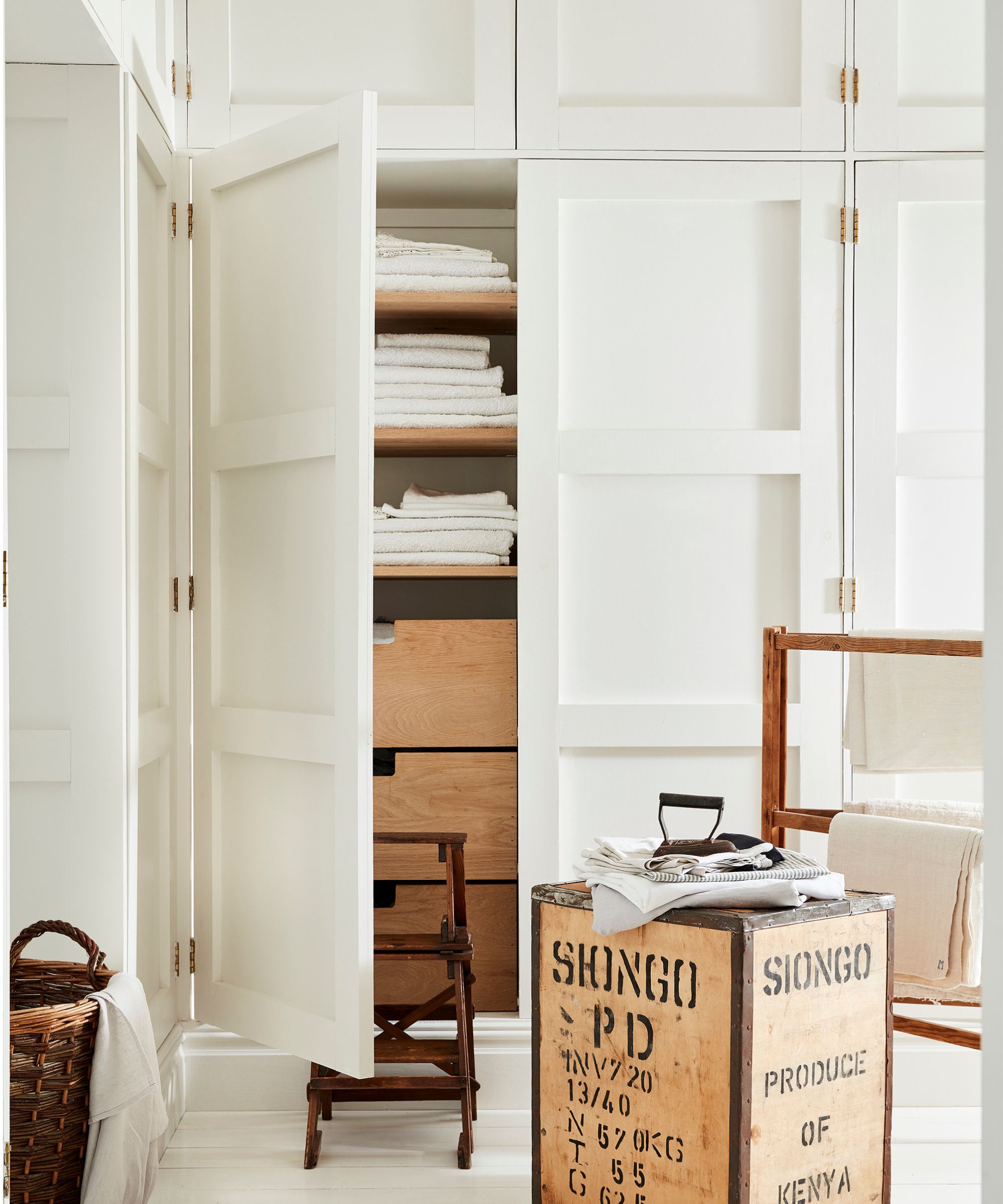 Placards blancs intégrés aux murs, une porte s'ouvrant révélant une armoire à linge remplie de serviettes et de literie, un repose-pieds en bois et un porte-serviettes à côté.