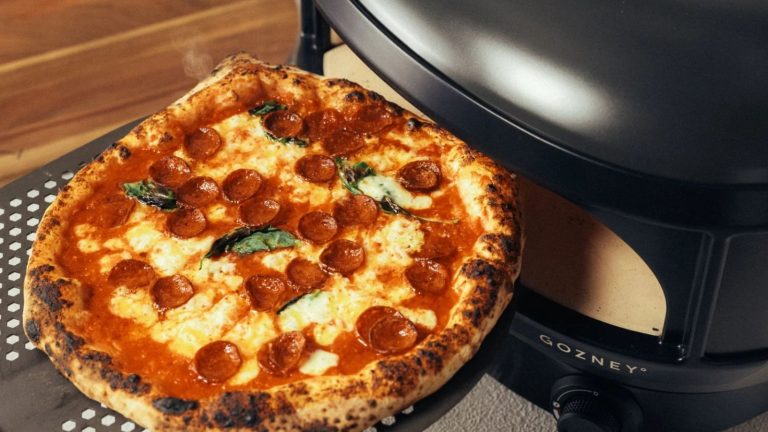 A cooked pepperoni pizza coming out of a black slimline pizza oven on a perforated pizza slice.