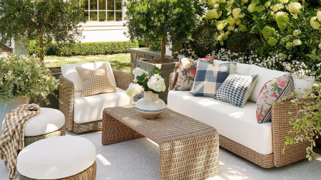 an outdoor living room with wicker furniture, white cushions, colorful pillows, a coffee table, a vase of flowers, two ottomans, and lots of greenery around the scene