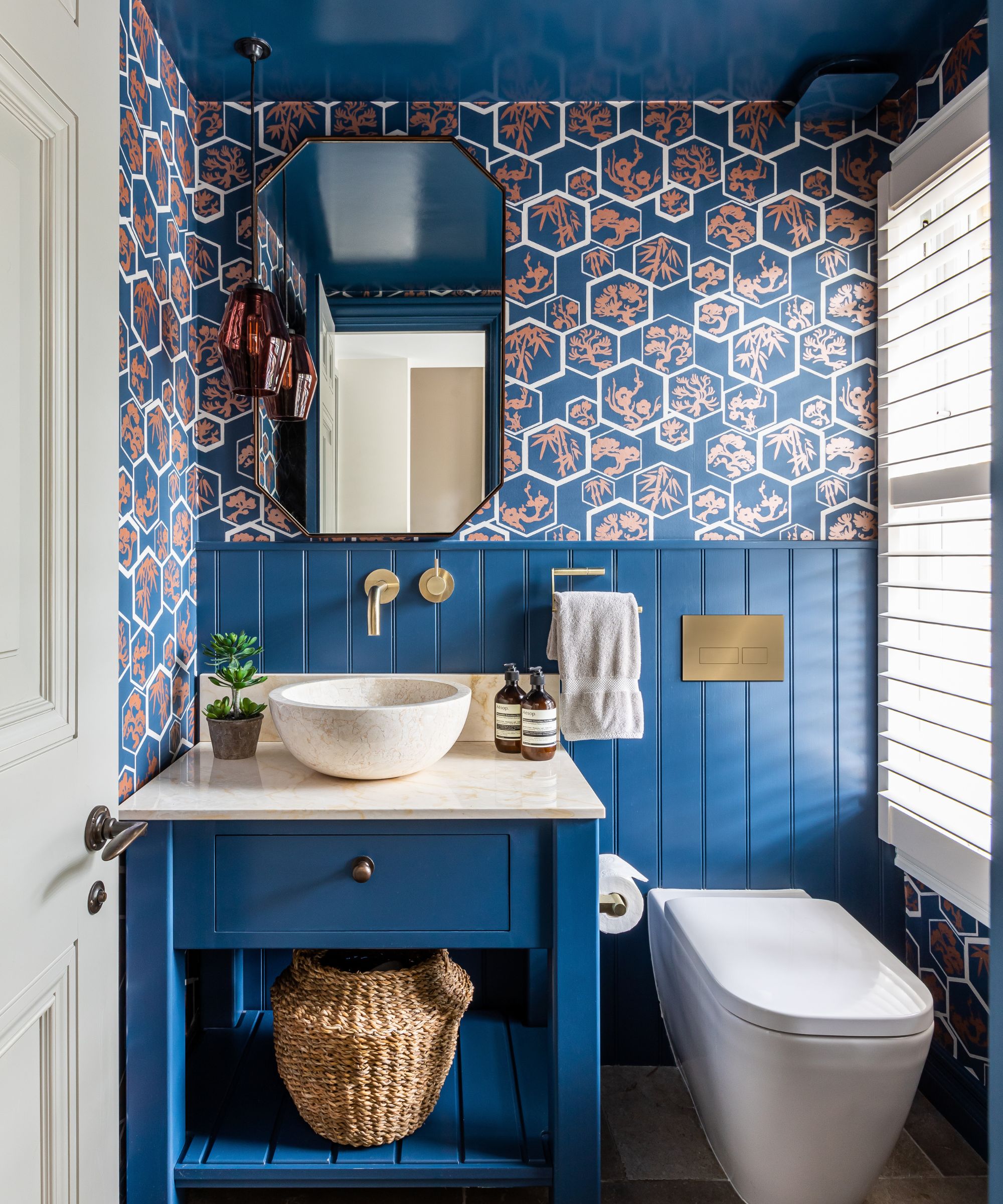 Salle de bains avec plafond bleu et boiseries peintes en bleu, meuble sous-vasque bleu avec comptoir et lavabo en marbre, papier peint sur la moitié supérieure du mur, longue fenêtre avec store blanc et quincaillerie dorée
