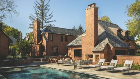 A luxury brick estate features a large swimming pool and a flagstone patio. The outdoor living area includes a massive brick fireplace with built-in wood storage, modern lounge chairs, and a sleek outdoor kitchen.