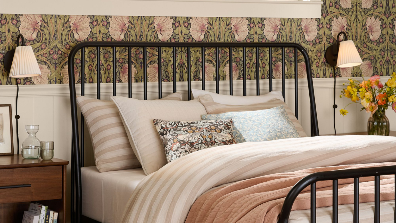 Pas de câblage, pas de problème – Les appliques enfichables transforment l’éclairage mural en un jeu d’enfant, et ces 12 sont les choix les plus chics a bedroom with floral wallpaper on the walls, a black metal bedframe, light pink bedding, and two black plug in wall sconces on either side of the bed