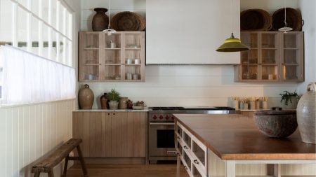 Neutral kitchen with large range cooker and wall mounted wooden cabinets with uplighting. There is a wooden island off center, to the front, and a narrow bench on the left with crittal white windows and translucent window treatments