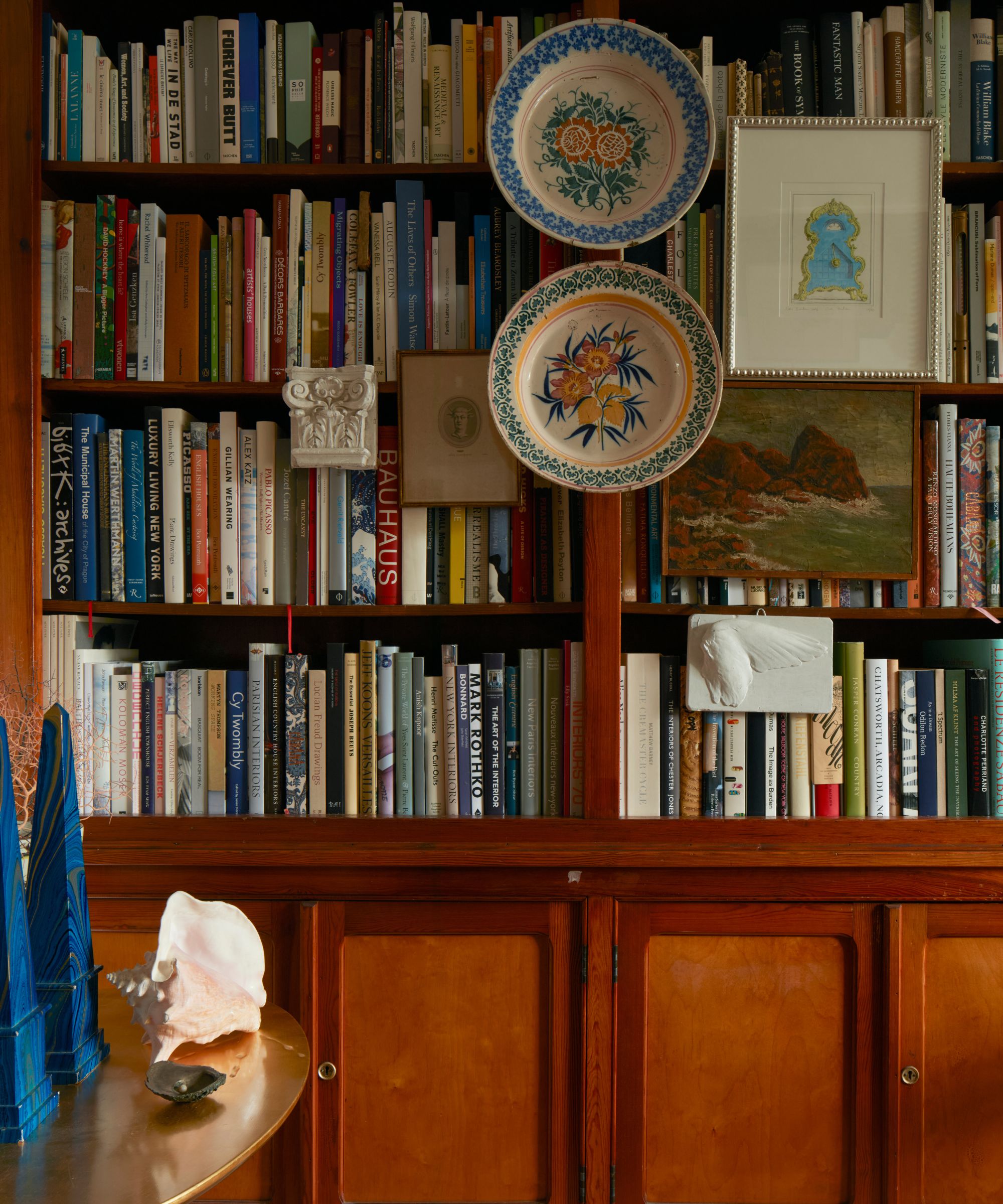 Bibliothèque en bois sombre avec de nombreux livres et assiettes en céramique accrochés devant, ainsi que des peintures et gravures vintage à côté d'une table ronde dorée avec un objet coquillage et des presse-papiers