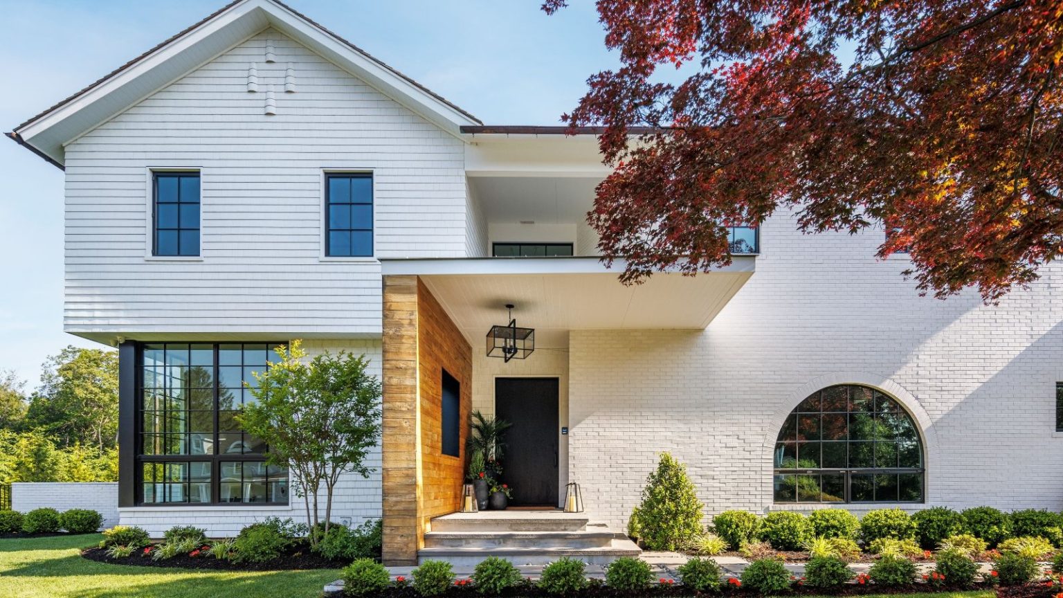 La réinitialisation de sécurité du printemps : 5 améliorations du porche pour un printemps plus sûr Bright, modern home exterior, with large windows and white walls. The front porch has a brick wall to the left side, and potted plants by the black front door. There are small bushes and a paved pathway in the front yard.