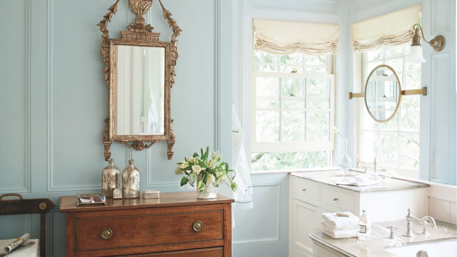 Benjamin Moore dévoile ses 4 couleurs de salle de bains les plus populaires – « Ces nuances adoptent une approche facile de la couleur, ce qui les rend intrinsèquement adaptables » Traditional bathroom with pale blue painted walls, mahogany sideboard with an ornate gold mirror above it, marble bathtub and vanity unit with sink, and two windows with cream roman blinds