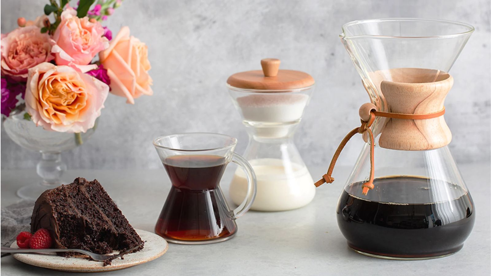 Une cafetière Chemex remplie de café noir, avec une tasse en verre remplie de café, un porte-lait en verre et une petite assiette de gâteau au chocolat. Derrière se trouve un vase en verre rempli de roses et le fond est gris.