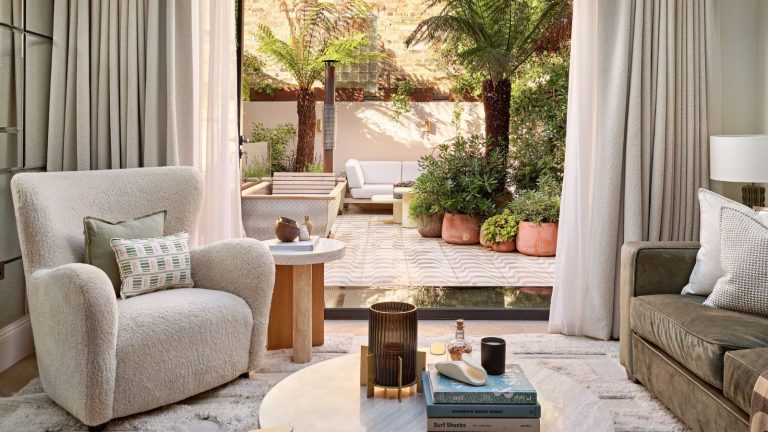 Fatigué des dégâts manqués ? L’aspirateur le plus vendu de Dyson vient d’atteindre son prix le plus bas de 2026 – Pourquoi ce professionnel ne jure que par lui Bright living room space with white rug, white plush armchair, and coffee table with books and glass ornament. A large open door is pictured in the background, revealing sunny outdoor space with plants and outdoor seating.