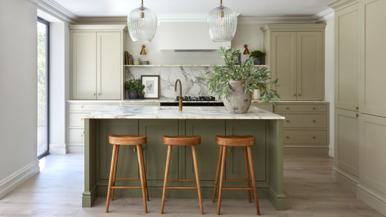 A kitchen with sage green cabinets and a darker green island. A large stone vase with green steps on the island beside a sink, and two glass pendants hanging from the ceiling above.