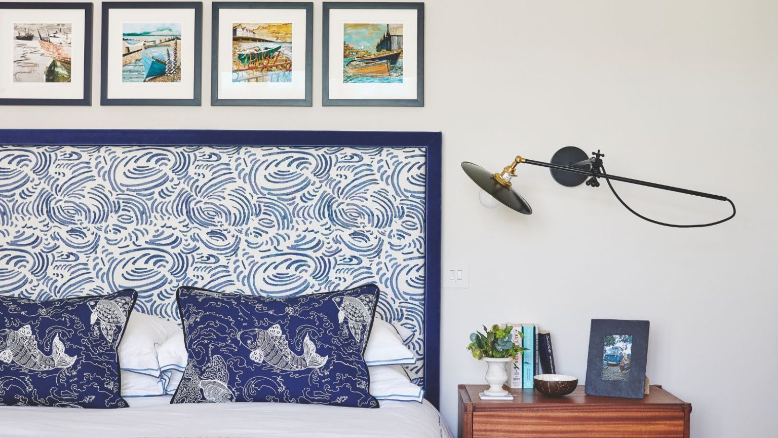 L’élément unique que les experts en Feng Shui vous conseillent de retirer de votre table de chevet en avril pour « favoriser un repos profond et une transition en douceur dans la journée » A white bedroom with double bed to the left with statement blue and white patterned headboard, and blue cushions patterned with koi fish. To the right of the headboard is a brown wood bedside table with a small plant, books, a small bowl, and a framed photograph. Above is a wall-mounted angle poise lamp, and above the bed are four small, square framed artworks.