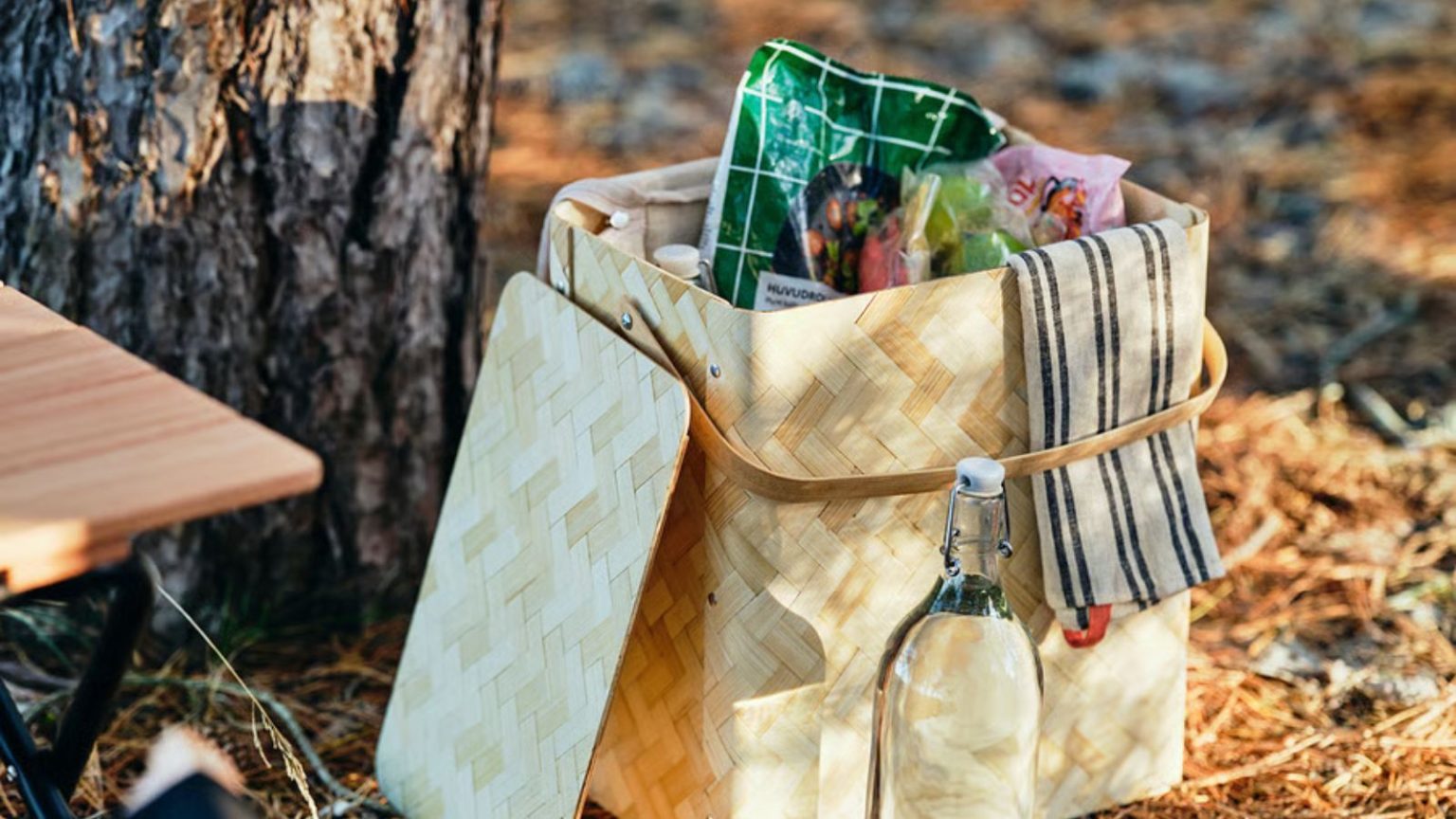 La glacière à panier en édition limitée d’IKEA est l’essentiel du pique-nique le plus chic de cet été – c’est la mise à niveau élégante de la glacière dont vous avez besoin A woven wooden basket with a cooler bag full of snacks inside, on a floor with fallen pine needles.