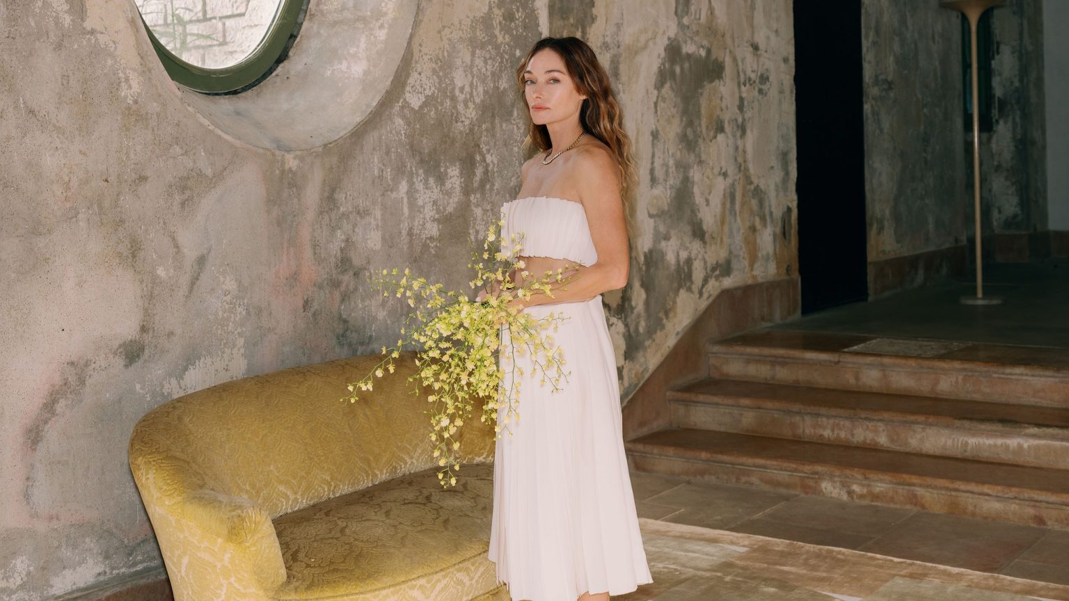« Passer au crible les déchets, c’est amusant » – La créatrice superstar Kelly Wearstler explique comment elle parvient toujours à trouver les trésors dans la poubelle Kelly Wearstler stands in a brutalist room holding a bouquet of yellow flowers