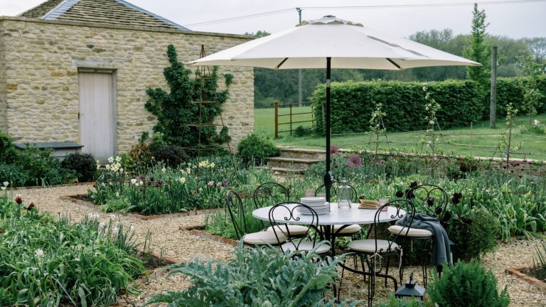 Cette option de siège simple peut donner à votre petit patio une atmosphère européenne élégante – Pourquoi les meubles de bistro sont le style à connaître en 2026 English country garden with flower beds, a brick outbuilding, a lawn looking out onto a field, and a round table with wrought iron bistro chairs and a parasol above