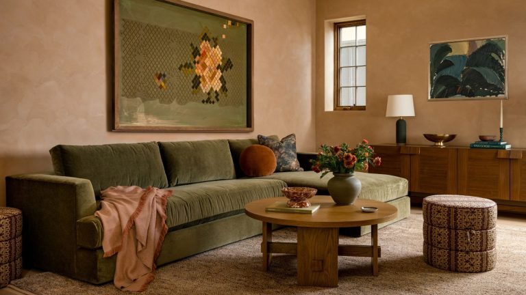 Votre salon n’a pas l’air plat à cause de votre palette de couleurs, c’est votre palette de textures qui est erronée. Voici comment superposer comme un designer pour ajouter de la profondeur à n’importe quelle pièce. Warm living room with plaster walls, green velvet sectional sofa, wooden coffee table, a floral ottoman, a fluffy rug and a large green and yellow painting of a flower above the sofa