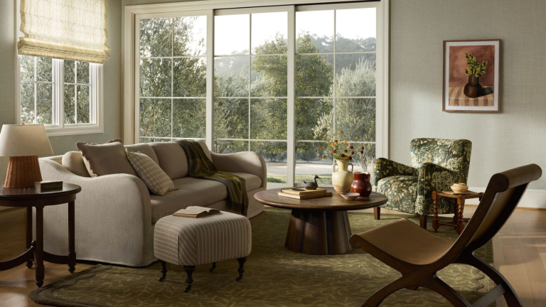 La nouvelle collection d’été de Joon Loloi allie l’artisanat traditionnel à l’ambiance du Sud – cela pourrait bien être la sortie la plus désirable de la marque à ce jour a living room with a taupe couch, a wooden coffee table, a brown sculptural chair, and a patterned chair. The room features a wall of windows and an artwork hanging from the wall