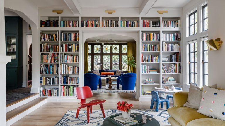 Les designers sont obsédés par le « Book Drenching » – la tendance 2026 qui enveloppe toute votre pièce de couleur et de caractère Living room with white painted walls, floor-to-ceiling bookcase with books, yellow velvet sofa, red chair on a rug, looking into another living room with blue armchairs and a blue sofa