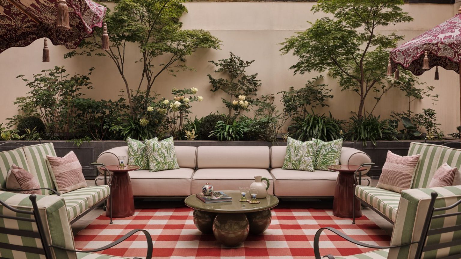 Pourquoi les designers « tapissent » leurs jardins en 2026 – « Nous avons passé si longtemps à adoucir nos intérieurs qu’il est logique que nos jardins emboîtent le pas » Garden with plants growing on the wall, a large red gingham rug with a large cream sofa on top, green metal armchairs with striped green cushions, and a marble green round coffee table at the centre.