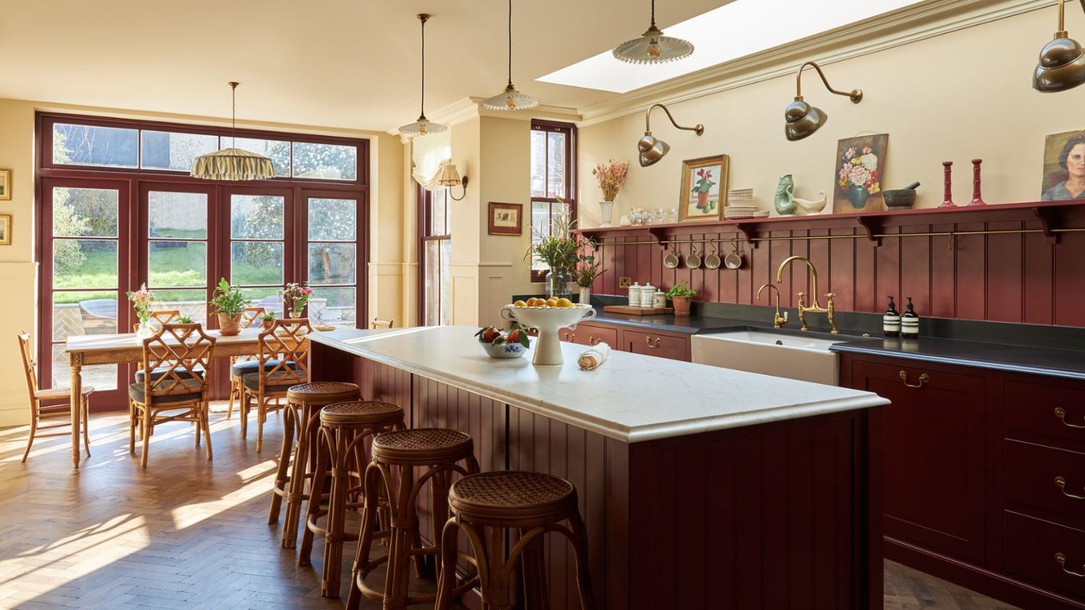 La couleur inattendue de la cuisine que nous pensions être une « phase » est désormais la plus grande source d’ambiance de 2026 A large kitchen with the cabinets and island painted oxblood red, a paneled backsplash, black countertops on the perimeter cabinets, and a white marble counter on the island.